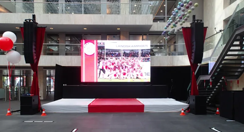 Laag podium met witte podiumrok en ingenaaide rode middenstrook, met een ledscherm met het Ajax-logo en de tekst Landskampioen